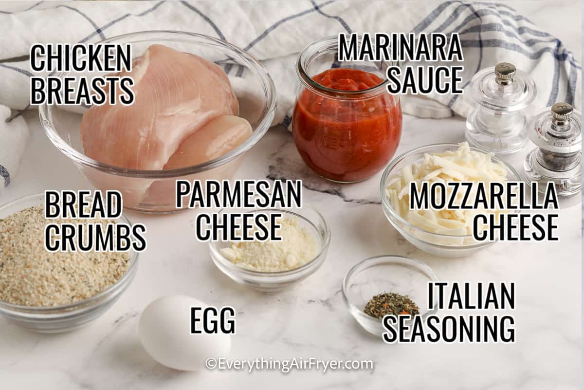 ingredients assembled to make chicken parmesan, including chicken breasts, marinara sauce, bread crumbs, parmesan cheese, mozzarella cheese, egg, and italian seasoning